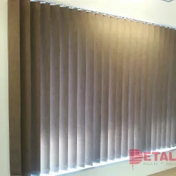 Beżowe pionowe panele okienne z tkaniny, zamontowane na białej szynie, z widocznym logo firmy w prawym dolnym rogu.