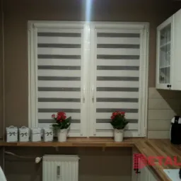 Nowoczesna kuchnia z oknem przesłoniętym roletami w poziome pasy, dekorowana doniczkami z czerwonymi kwiatami i ozdobnymi pudełkami.