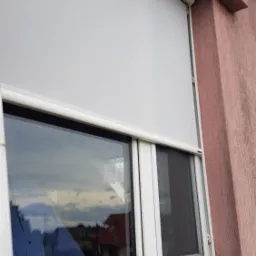 Okna z białą ramą i częściowo opuszczoną białą roletą na tle różowej ściany zewnętrznej, widoczna także czarna moskitiera w oknie.