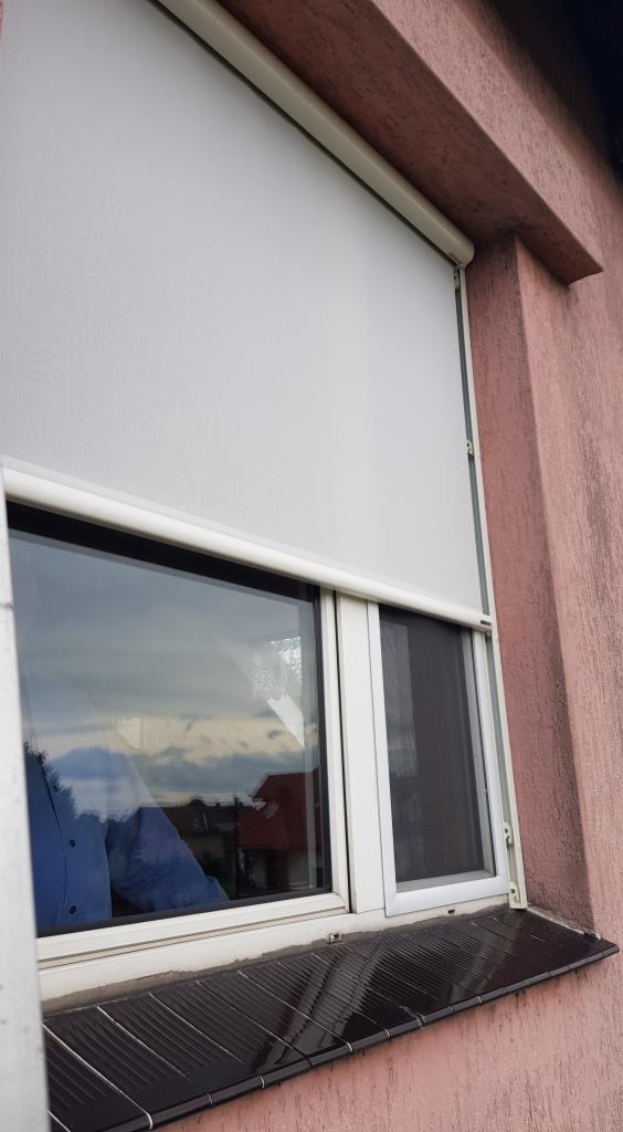 Okna z białą ramą i częściowo opuszczoną białą roletą na tle różowej ściany zewnętrznej, widoczna także czarna moskitiera w oknie.