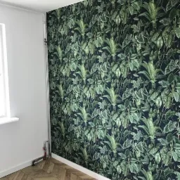 Ściana wykończona tapetą w tropikalne liście, jasna podłoga ułożona w jodełkę, widoczne rury grzewcze przy ścianie.