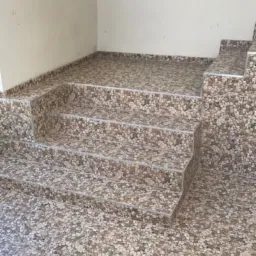 Schody tarasowe wyłożone płytkami imitującymi kamień, z widoczną hydroizolacją pod kaflami, zabezpieczającą przed warunkami atmosferycznymi; jasne wnętrze z oknem w tle.