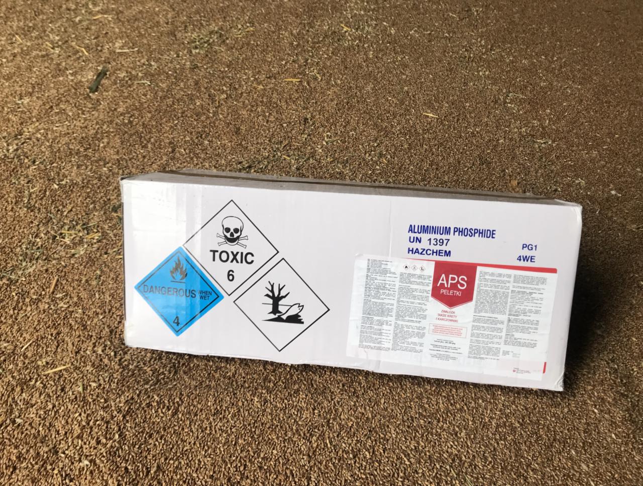Białe pudełko z ostrzegawczymi symbolami toksyczności i substancji niebezpiecznych, zawierające fosforek glinu, leżące na brązowym podłożu.