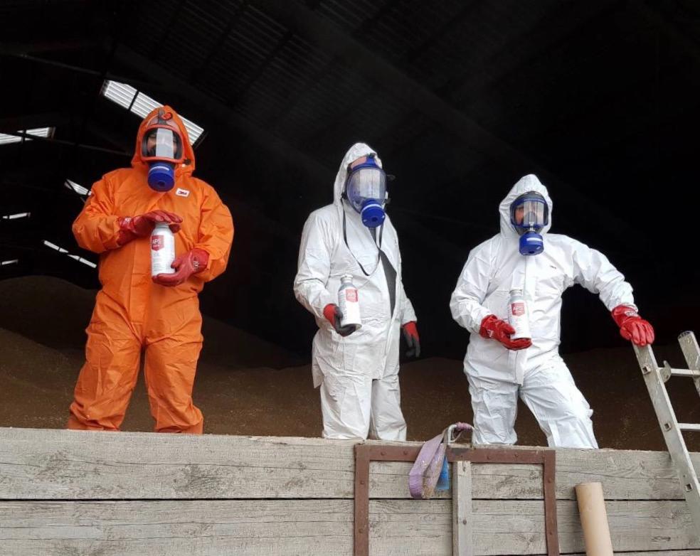 Trzech mężczyzn w kombinezonach ochronnych i maskach gazowych trzymających białe butelki na tle sterty ziarna w magazynie.