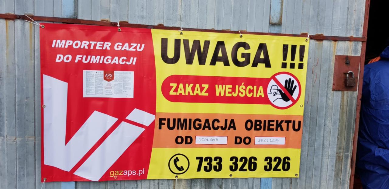 Baner informujący o fumigacji obiektu z datami i numerem telefonu, zawieszony na metalowej ścianie, obok widoczna osoba w kombinezonie ochronnym.
