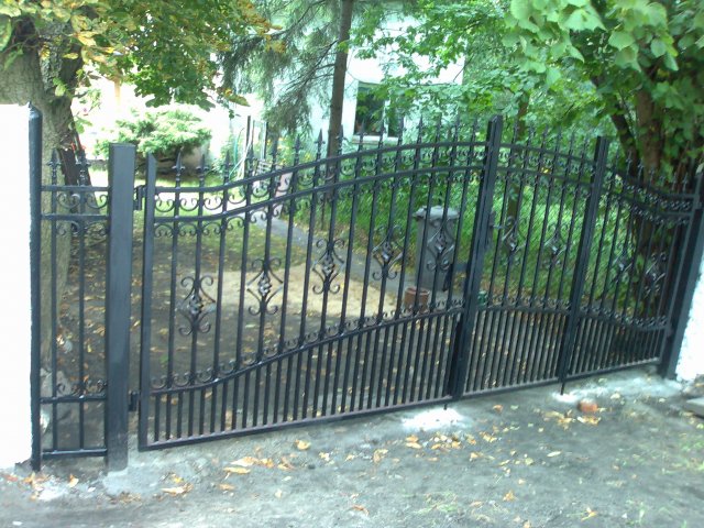 Dwuskrzydłowa, czarna brama wjazdowa z kutymi zdobieniami, osadzona na betonowych słupkach, prowadząca na posesję z zielenią w tle.