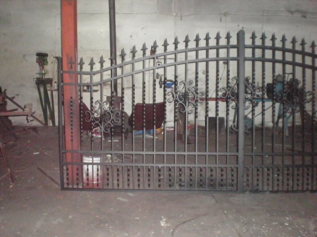 Czarna, metalowa brama z dekoracyjnymi elementami w warsztacie, widoczny sprzęt spawalniczy i narzędzia w tle.