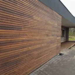 Nowoczesny dom z elewacją z poziomo ułożonych desek drewnianych, szara ściana i kostka brukowa na pierwszym planie, widoczny fragment tarasu i elementy ogrodu w tle.