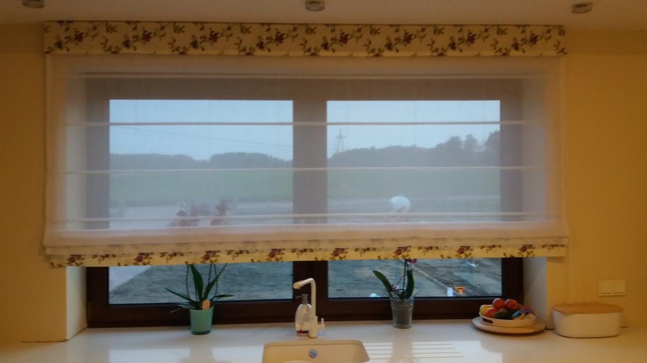 Zasłona rzymska z motywem kwiatowym w kuchni, widok zza okna na zewnątrz, roślinki w doniczkach na parapecie.