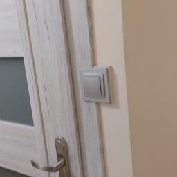 Łukasz Woznica - Drzwi wewnętrzne z szarą okleiną imitującą drewno, srebrna klamka z zamkiem i szary włącznik światła na beżowej ścianie.