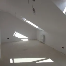 Poddasze w trakcie remontu, widoczne okno dachowe, białe ściany z otworami po gniazdkach elektrycznych, surowa podłoga, grzejnik i wisząca żarówka na kablu.