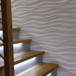 Drewniane schody z podświetleniem LED, przylegające do ściany z dekoracyjnymi, falistymi panelami 3D, widok z dołu.