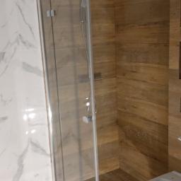 Łukasz Woznica - Nowoczesna kabina prysznicowa z drewnopodobnymi płytkami na ścianach, szklane drzwi z chromowanymi zawiasami, biała wisząca toaleta, marmurowe płytki podłogowe.