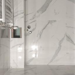 Łukasz Woznica - Biała łazienka z marmurowymi płytkami na ścianach i podłodze, widoczny grzejnik drabinkowy po lewej stronie oraz przygotowane otwory na armaturę.