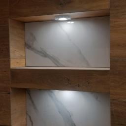 Łukasz Woznica - Wnęki w ścianie wykończone drewnopodobnymi płytkami i białym marmurem, oświetlone punktowymi lampkami LED, z widocznym przyciskiem spłuczki i klapą rewizyjną w dolnej części.