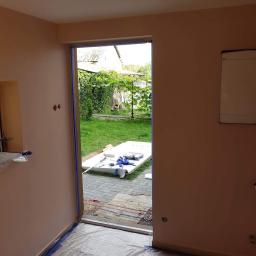 Łukasz Woznica - Przygotowania do malowania ścian w pomieszczeniu z widokiem na ogród: ściany pomalowane na jasny róż, zabezpieczone folią podłogi i taśmą malarską framugi drzwi, przez które widać zielony trawnik...