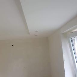 Łukasz Woznica - Świeżo pomalowane wnętrze z gładką, białą ścianą i sufitem z zamontowanymi dwoma punktowymi lampami LED. Widoczne ślady po instalacji elektrycznej na ścianie.