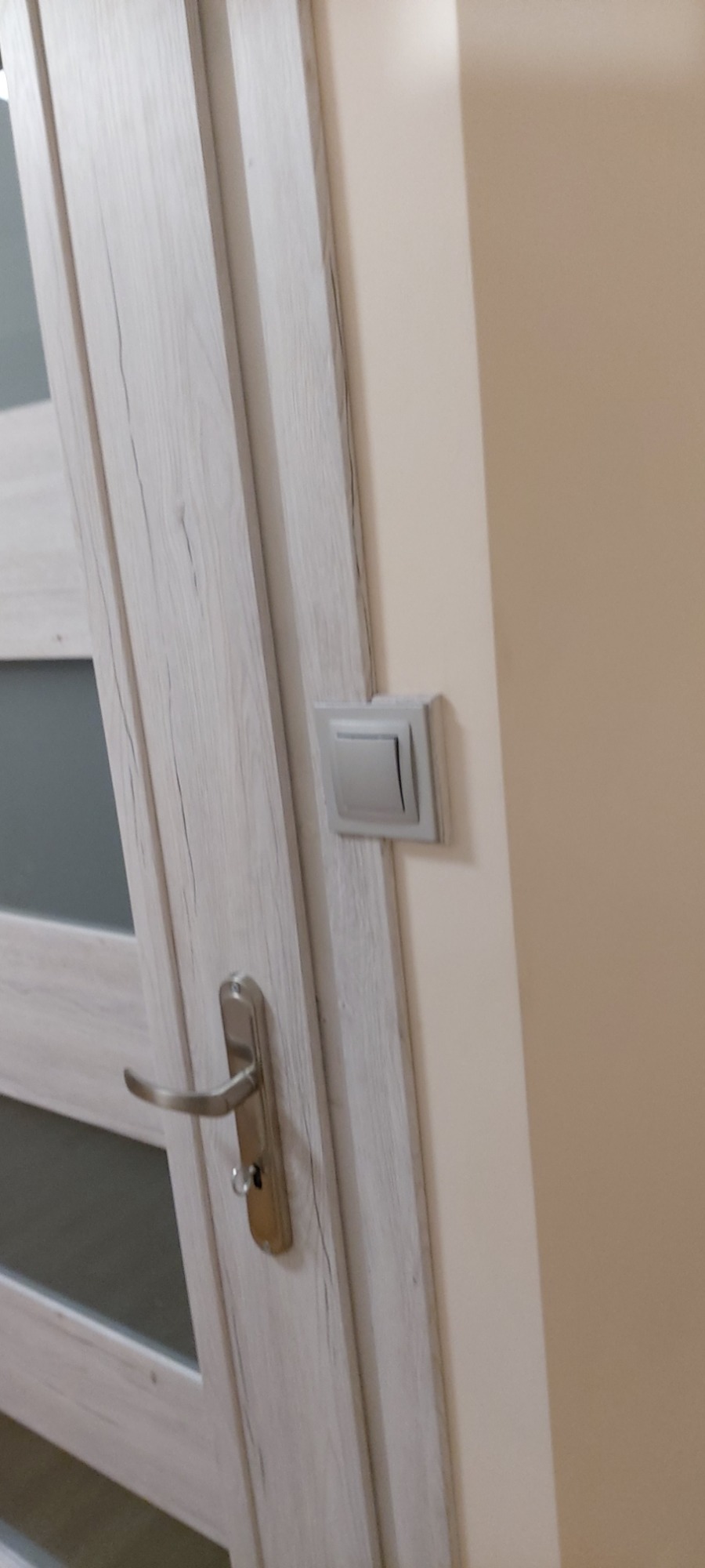 Drzwi wewnętrzne z szarą okleiną imitującą drewno, srebrna klamka z zamkiem i szary włącznik światła na beżowej ścianie.