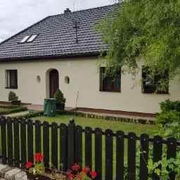 Dom jednorodzinny z beżową elewacją, ciemną dachówką i drewnianymi oknami, widoczny zza ciemnego płotu z kwiatami, w tle zielony trawnik i krzewy.