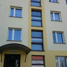 Fasada bloku mieszkalnego z żółtą elewacją, widoczne okna z brązowymi ramami oraz wejście do budynku z numerem 13a.