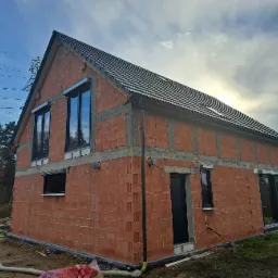 Budynek w stanie surowym otwartym z zamontowanymi oknami i roletami zewnętrznymi, widoczne pustaki ceramiczne, betonowe nadproża, czarne rynny i dachówka, w tle drzewa i pole.