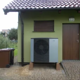 Szara pompa ciepła typu monoblok zamontowana na betonowych wspornikach przed zielonym domem z brązowymi akcentami, widoczne dwa brązowe pojemniki na śmieci.