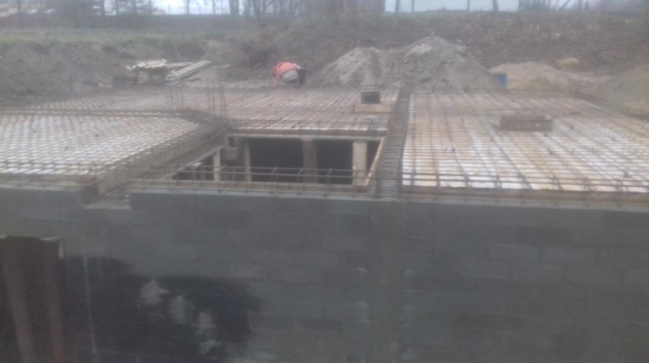 Wykopany fundament domu murowanego z widoczną konstrukcją zbrojeniową i betonowymi ścianami, z betoniarką w tle i ziemią.