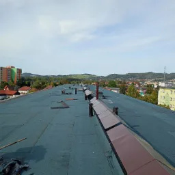 Płaski dach pokryty papą termozgrzewalną z elementami wentylacyjnymi i obróbkami blacharskimi, widok z góry na tle zabudowy miejskiej i wzgórz.