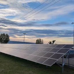 Rząd paneli słonecznych na trawie, z ciężarówką i samochodami w tle, pod wieczornym niebem z chmurami.