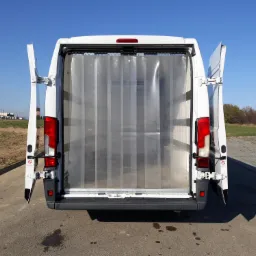 Wnętrze białego busa z otwartymi drzwiami i kurtyną paskową, widoczne z tyłu pojazdu, na zewnątrz asfalt i zieleń