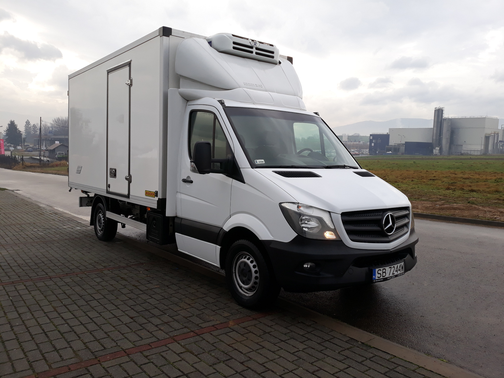 Biały samochód dostawczy Mercedes-Benz z chłodnią na tle przemysłowym i pochmurnego nieba, zaparkowany na brukowanym chodniku.