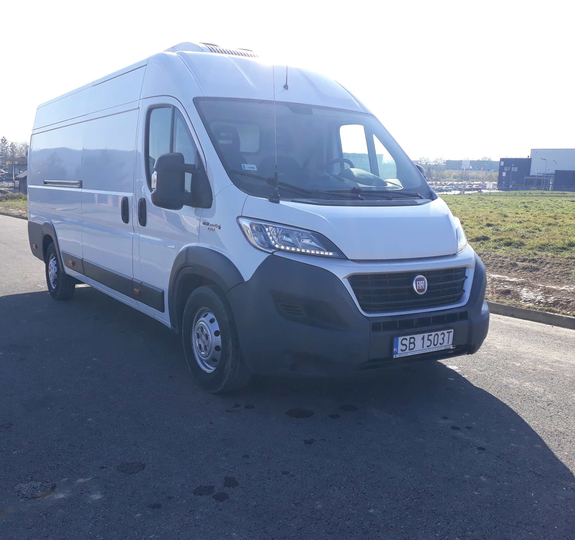 Biały samochód dostawczy Fiat Ducato z widoczną tablicą rejestracyjną SB 1503T, zaparkowany na asfaltowej drodze z polem w tle.