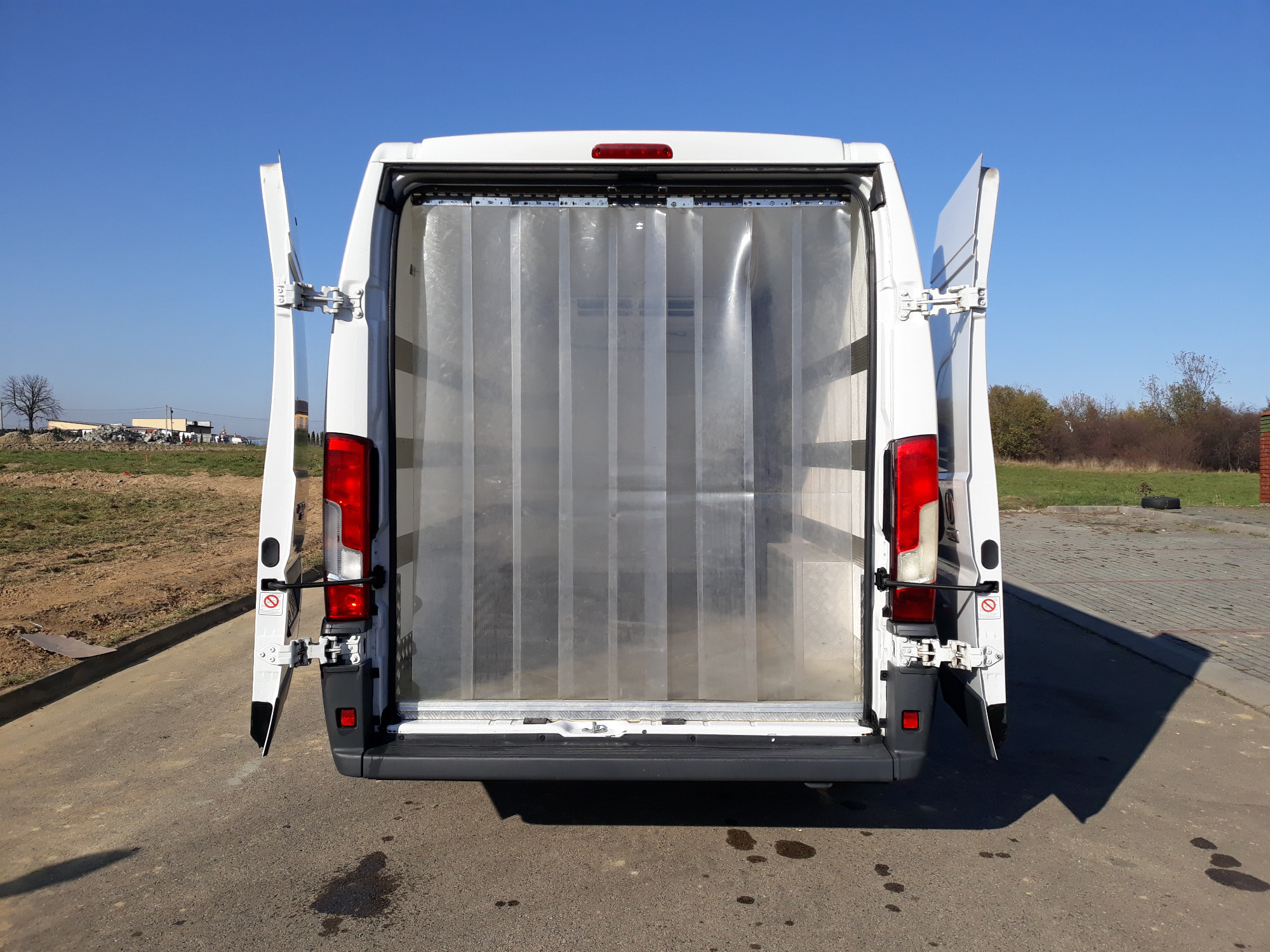 Wnętrze białego busa z otwartymi drzwiami i kurtyną paskową, widoczne z tyłu pojazdu, na zewnątrz asfalt i zieleń