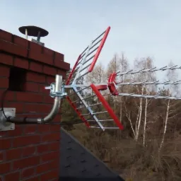 Czerwono-srebrna antena telewizyjna zamontowana na ceglanym kominie, widok z dołu na tle drzew.