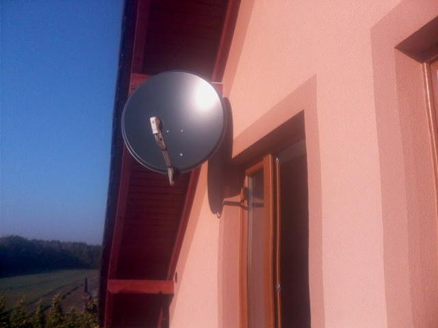 Szara antena satelitarna zamontowana na elewacji budynku w pobliżu okna z drewnianą ramą, widok na zielone pola w tle.