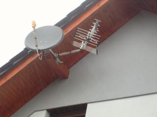 Antena satelitarna i antena naziemna zamontowane na drewnianej podbitce dachu, widoczne z boku na tle białej ściany budynku.