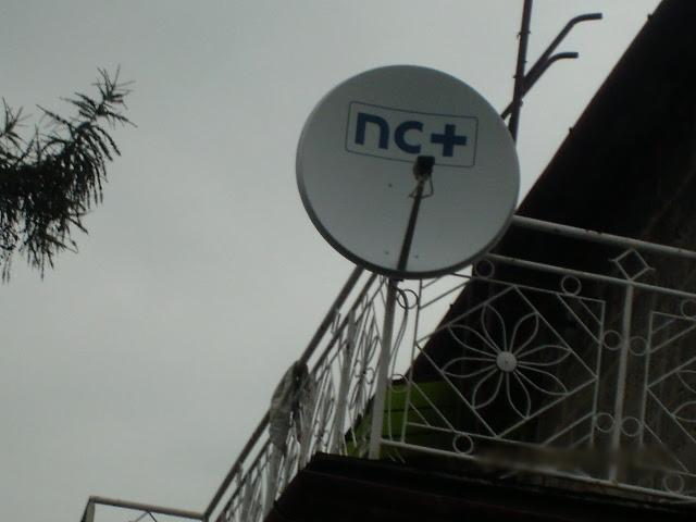 Biała antena satelitarna z logo nc+ zamontowana na balkonie z ozdobną, białą balustradą, widok z dołu na tle pochmurnego nieba.