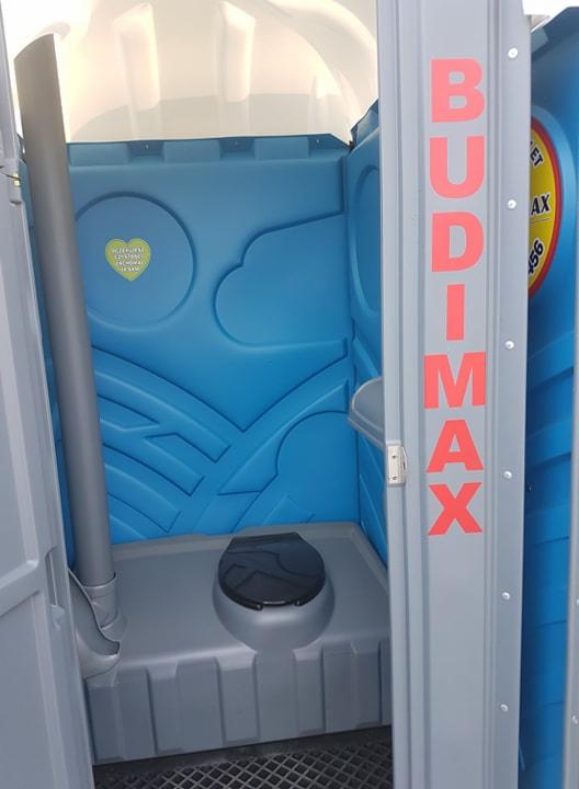 Wnętrze przenośnej toalety z niebieskimi ścianami, z widocznym logo firmy BUDIMAX na drzwiach i naklejką w kształcie serca na ścianie.