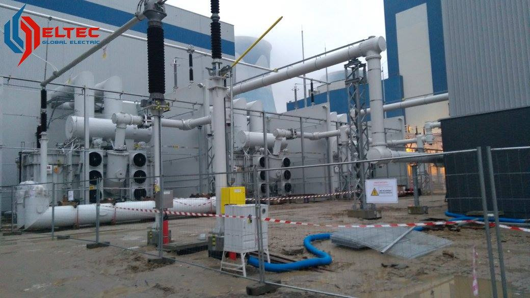 Elektrownia Opole: Rozbudowana infrastruktura elektryczna na zewnątrz budynku, widoczne transformatory, rury, kable oraz elementy zabezpieczające teren budowy, takie jak taśmy ostrzegawcze i barierki.