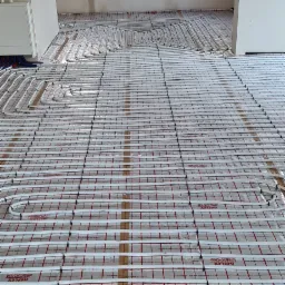 Instalacja ogrzewania podłogowego w nowym budynku: białe rurki układane na folii z nadrukiem, widok na surową ścianę z oknami.