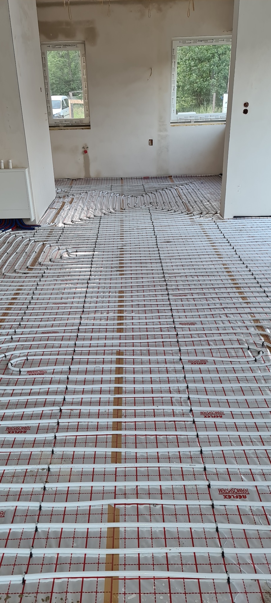 Instalacja ogrzewania podłogowego w nowym budynku: białe rurki układane na folii z nadrukiem, widok na surową ścianę z oknami.