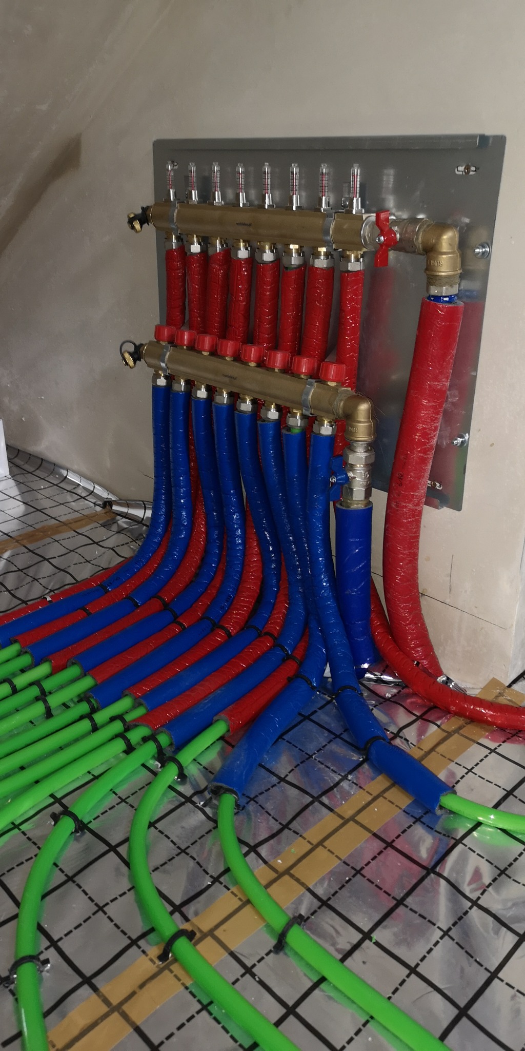 Rozdzielacz ogrzewania podłogowego z rurami w kolorach: czerwonym, niebieskim i zielonym, ułożonymi na folii izolacyjnej z nadrukowaną siatką.