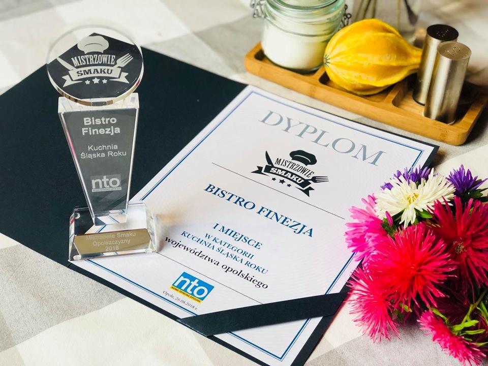 Nagroda i dyplom dla Bistro Finezja za I miejsce w kategorii Kuchnia Śląska Roku województwa opolskiego, obok bukiet kwiatów, solniczka i pieprzniczka oraz słoik z zawartością na drewnianej tacy.