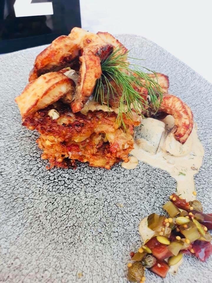 Wykwintnie podane danie: warstwowe placki ziemniaczane z chrupiącymi krewetkami, sosem śmietanowym i piklami na szarym talerzu o porowatej strukturze.