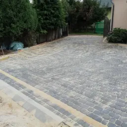 Ułożona szara kostka brukowa na podjeździe, z jasnymi pasami i obrzeżami, widoczne narzędzia pomiarowe oraz dom w tle.