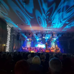 Widok na scenę z orkiestrą symfoniczną podczas koncertu, z widownią w tle i efektami świetlnymi na suficie.