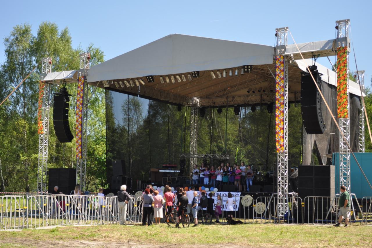 Scena plenerowa z oświetleniem i nagłośnieniem podczas występu chóru, widoczni widzowie za barierkami.