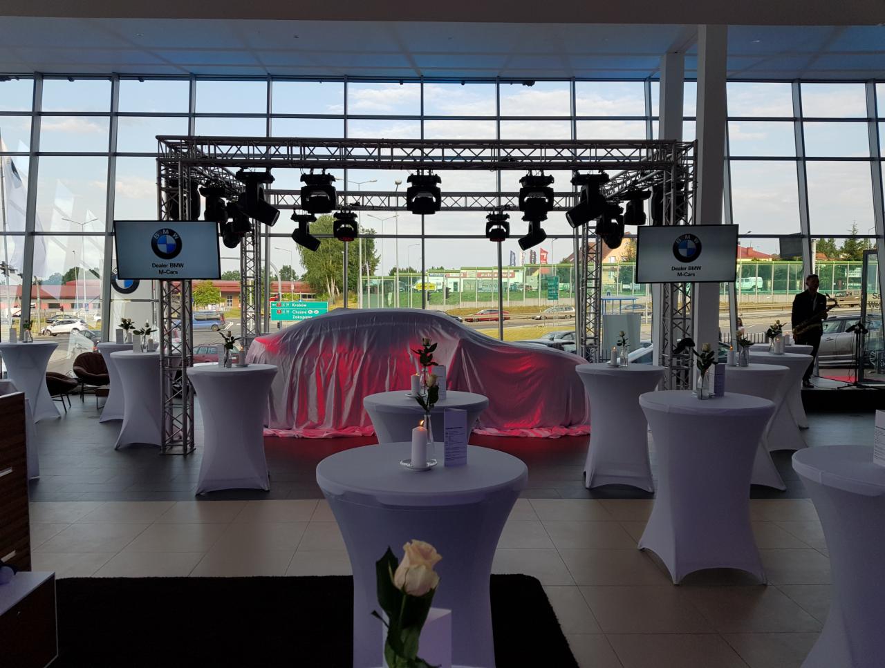 Prezentacja samochodu BMW M-Cars pod czerwoną płachtą, oświetlenie sceniczne, ekrany z logo BMW, białe stoliki koktajlowe z kwiatami w salonie samochodowym.