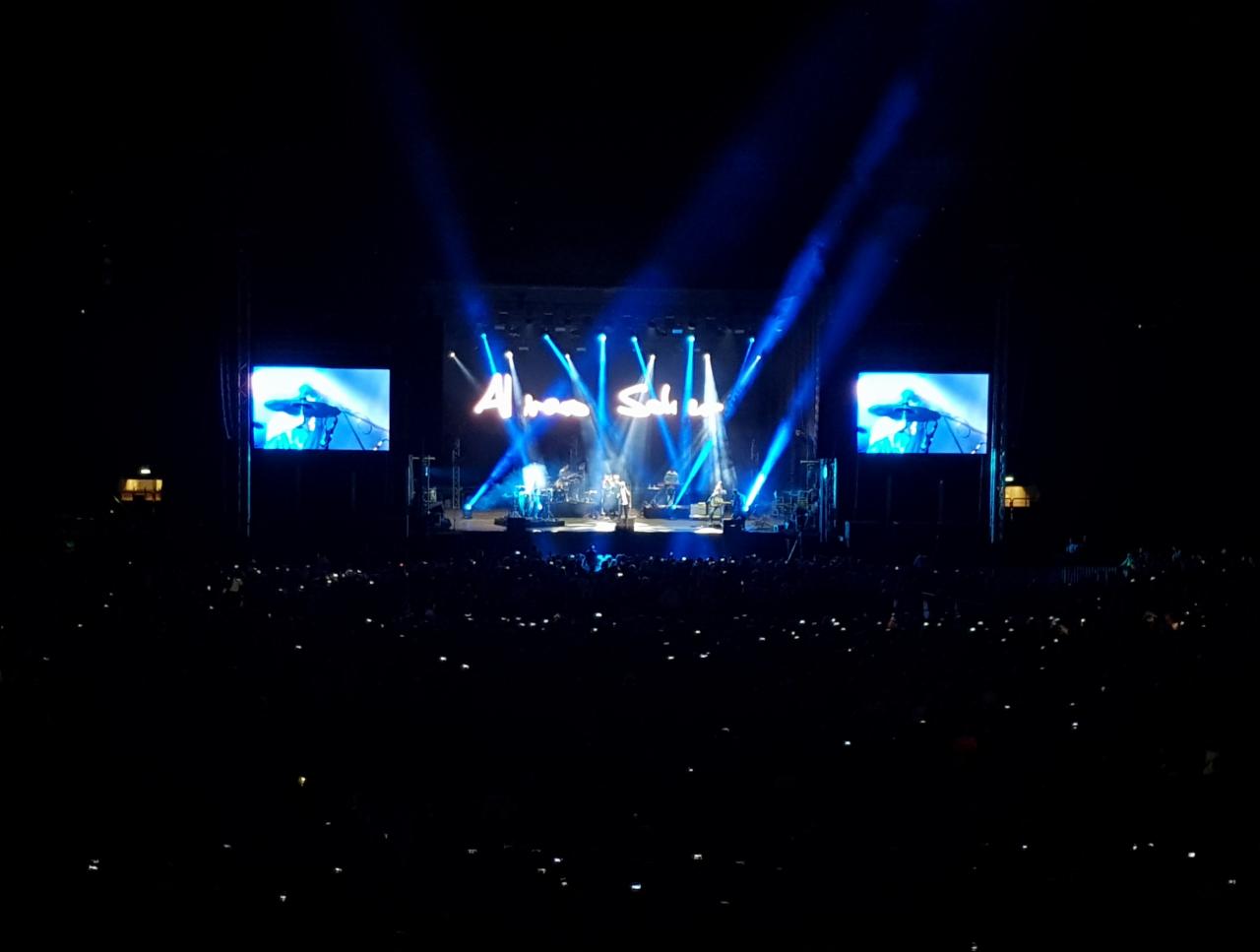 Koncert na dużej scenie z efektami świetlnymi i telebimami po bokach, widownia z podniesionymi telefonami.