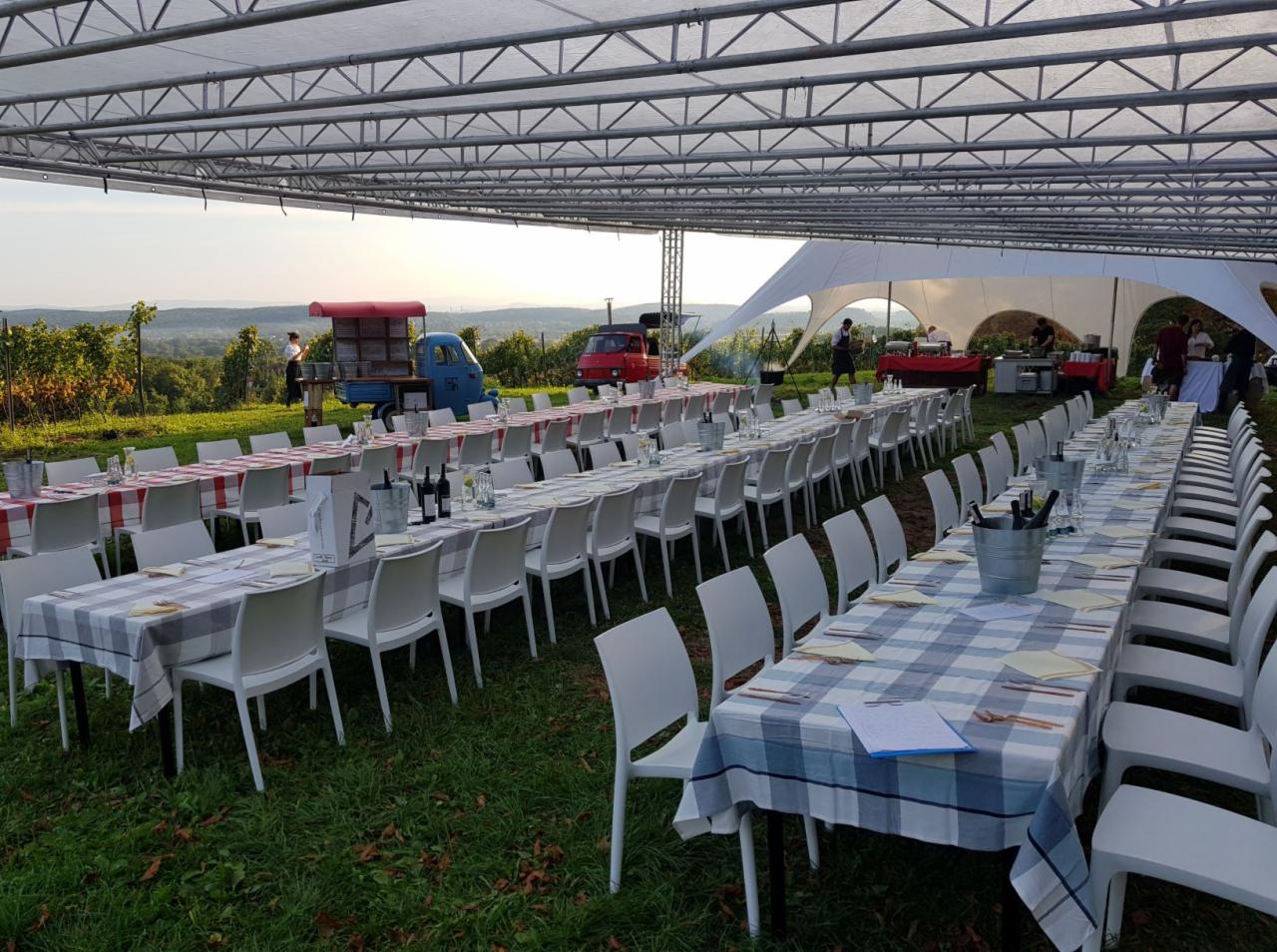 Długie stoły nakryte w kratę, ustawione na zewnątrz pod białym namiotem, przygotowane na przyjęcie z widokiem na wzgórza; w tle food trucki.
